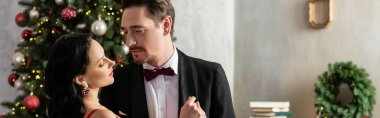 Takım elbiseli yakışıklı adam Noel ağacının yanında kırmızı şık elbiseli güzel kadını baştan çıkarıyor.