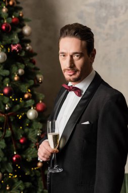 Smokinli yakışıklı zengin adam elinde şampanya bardağıyla evde süslü Noel ağacının yanında.