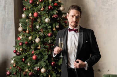 Smokinli, papyonlu, şampanya bardağıyla süslü Noel ağacının yanında duran zengin bir beyefendi.