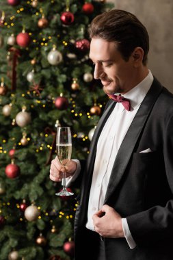 pleased wealthy man in tuxedo with bow tie holding champagne glass near decorated Christmas tree clipart