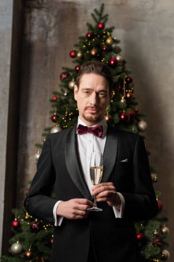 Takım elbiseli, papyonlu, şampanya bardağıyla süslü Noel ağacı taşıyan zengin bir beyefendi.
