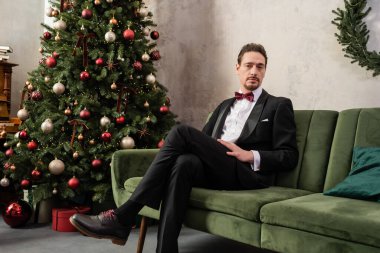İyi giyimli, sakallı, smokinli, papyonlu, Noel ağacının yanındaki koltukta oturan zengin bir adam.