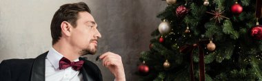Sakallı, smokinli, papyonlu iyi giyimli bir beyefendi Noel ağacına bakıyor.