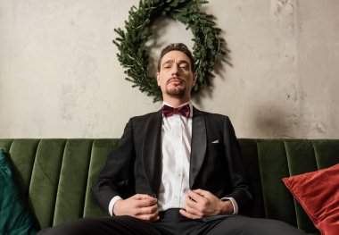 Sakallı, smokinli, papyonlu zengin bir beyefendi Noel çelenginin yanındaki koltukta oturuyor.