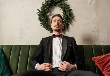 wealthy gentleman with beard wearing tuxedo with bow tie sitting on sofa near Christmas wreath clipart