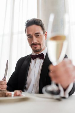 wealthy man in tuxedo cheering glass of champagne near delicious beef steak and looking at camera clipart
