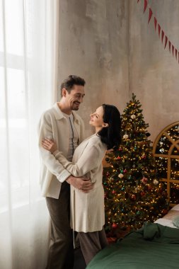 Evde mutlu bir çift gülümsüyor ve yatak odasındaki süslü Noel ağacının yanında sarılıyorlar.