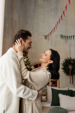 Evdeki neşeli kadın yatak odasındaki Noel ağacının yanında kocasını kucaklıyor.