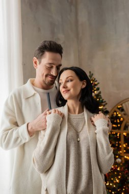 Rahat evdeki neşeli adam karısını kucaklıyor ve yatak odasındaki Noel ağacının yanında gülümsüyor.
