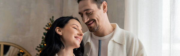 cheerful man hugging happy wife in cozy home wear near blurred Christmas tree, happy marriage banner