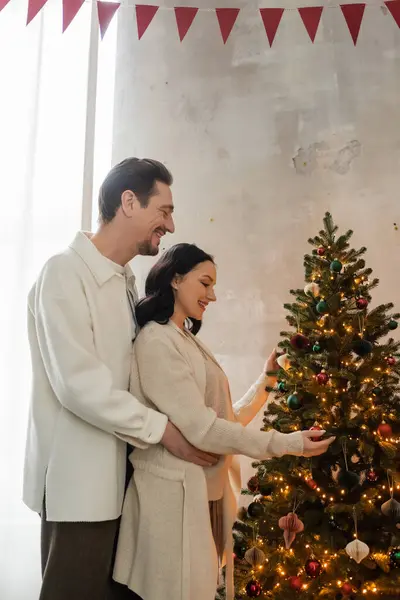 happily married couple in cozy home wear decorating Christmas tree in modern apartment, festive day
