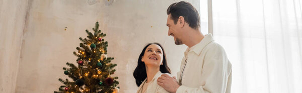 happily married couple in soft home wear decorating Christmas tree in modern apartment, banner