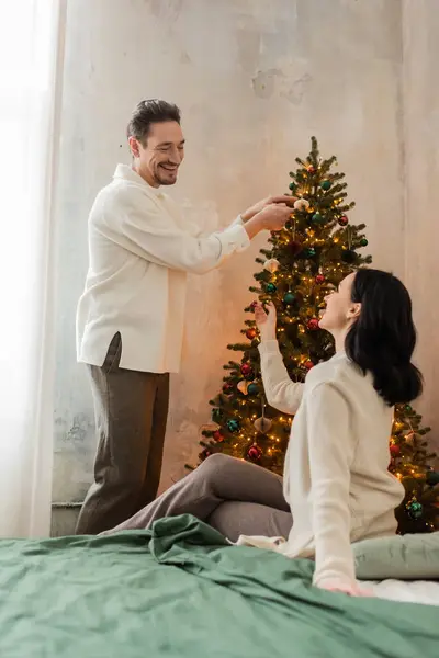 Homem Alegre Decorando Árvore Natal Olhando Para Esposa Morena Sentada — Fotografia de Stock