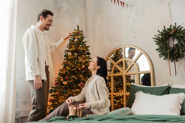 happy man decorating Christmas tree near wife wrapping present and sitting on bed, season of joy