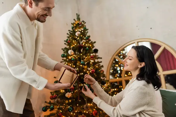 happily married couple holding gift box and ribbon, wrapping present near decorated Christmas tree