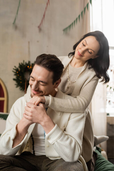 cheerful married couple holding hands, sitting on bed near Christmas wreath in modern apartment