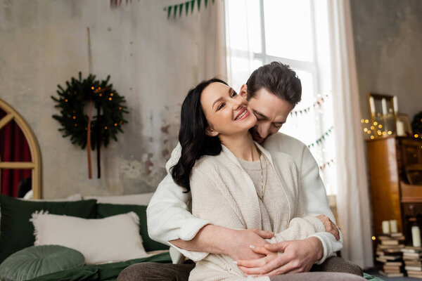 husband embracing joyful wife and sitting together on bed near blurred Christmas wreath on wall