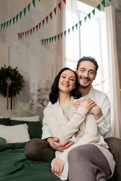husband embracing cheerful wife and sitting together on bed near blurred Christmas wreath on wall