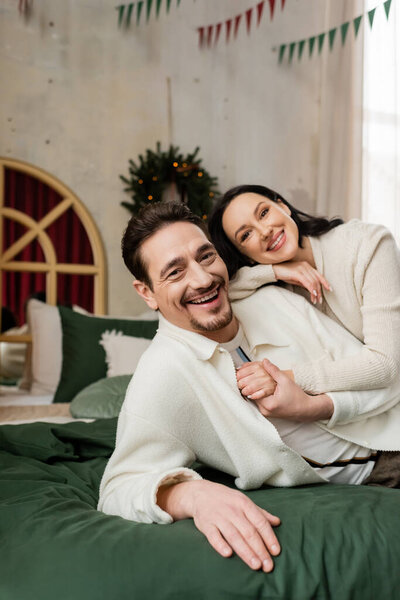 husband embracing cheerful wife and resting together on bed near blurred Christmas wreath on wall