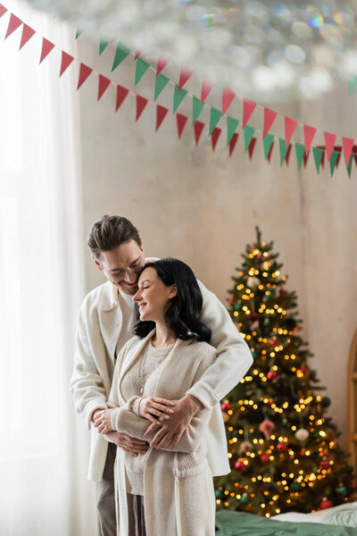 cheerful and married couple in home wear hugging and standing together near blurred Christmas tree