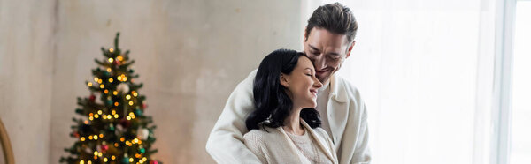 joyful married couple in home wear hugging and standing together near blurred Christmas tree, banner