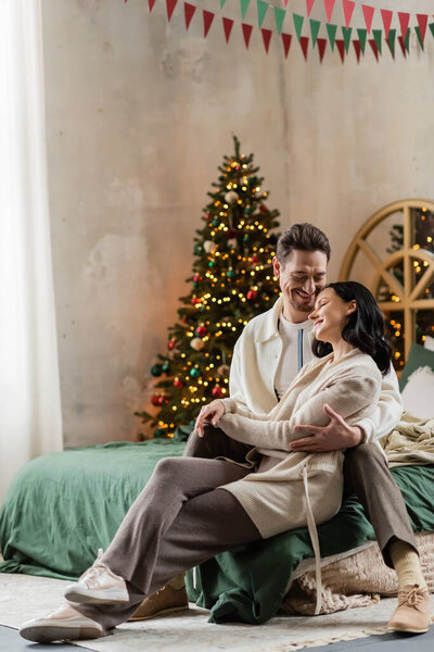 cheerful couple in home wear sitting on bed and holding hands near blurred Christmas tree at home