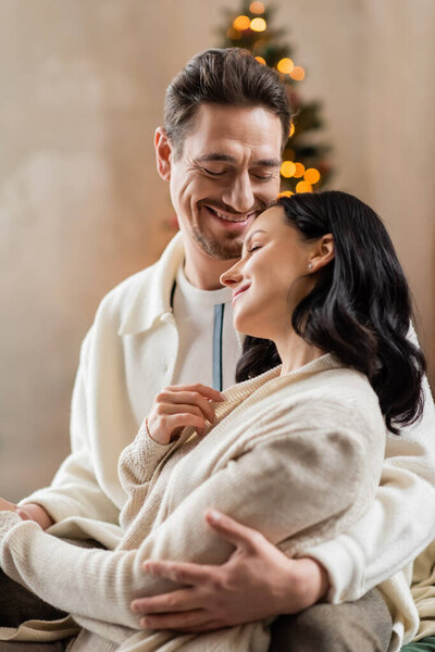 cozy living, happily married couple hugging each other near blurred Christmas lights on backdrop