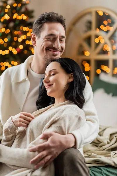 cozy living, happily married couple embracing each other near blurred Christmas lights on backdrop