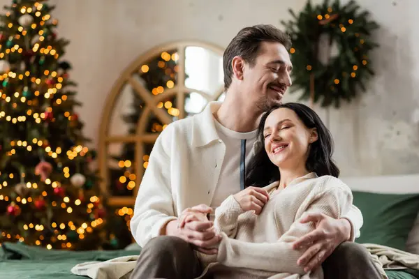 happily married couple embracing each other near blurred lights of Christmas tree on backdrop, cozy