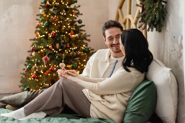 cheerful married couple spending cozy morning near blurred lights of Christmas tree on backdrop