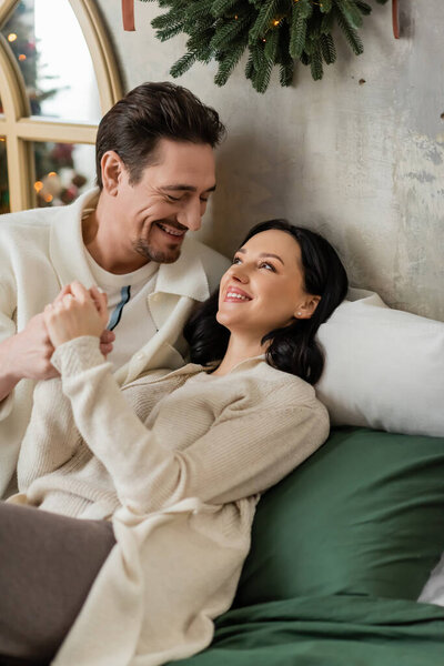 bearded man spending time together with happy wife and lying on bed near Christmas wreath on wall