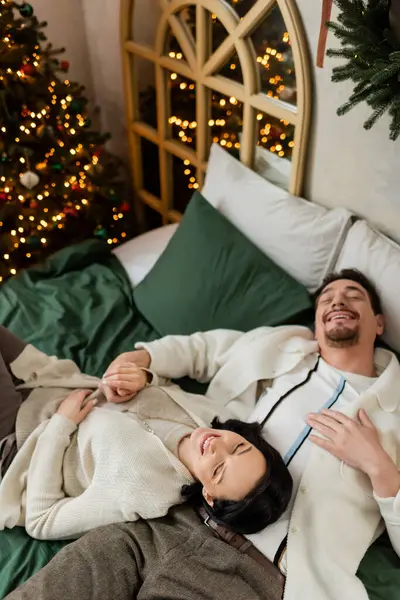 Stock image overhead view of joyful married couple spending cozy morning in bed near decorated Christmas tree