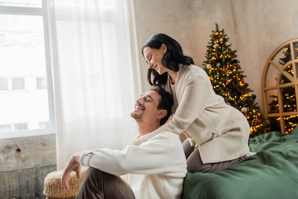 brunette woman hugging with husband and spending time together in bedroom near Christmas tree
