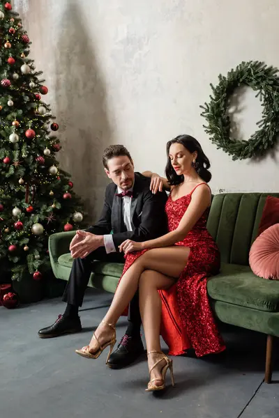 elegant and wealthy couple in evening attire sitting on sofa near decorated Christmas tree