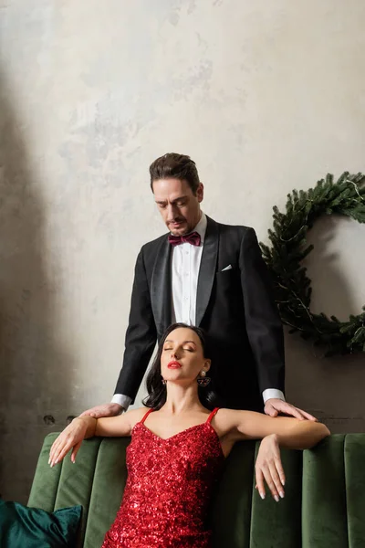 穿着燕尾服的英俊绅士看着穿着红衣服坐在沙发上的漂亮女人 富有的夫妇 — 图库照片