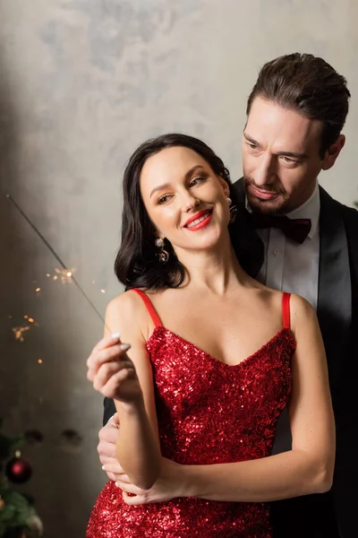 beautiful wealthy couple in formal attire smiling and looking at bright sparkler on Christmas eve
