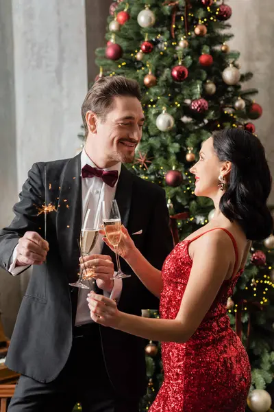 joyful married couple in formal attire holding champagne glasses and sparklers near Christmas tree