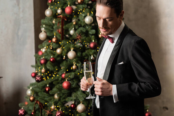 красивый богатый мужчина в смокинге с галстуком-бабочкой держа бокал шампанского рядом с украшенной елкой