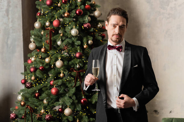 wealthy gentleman in tuxedo with bow tie holding champagne glass near decorated Christmas tree