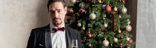 wealthy man in tuxedo with bow tie holding champagne glass near decorated Christmas tree, banner