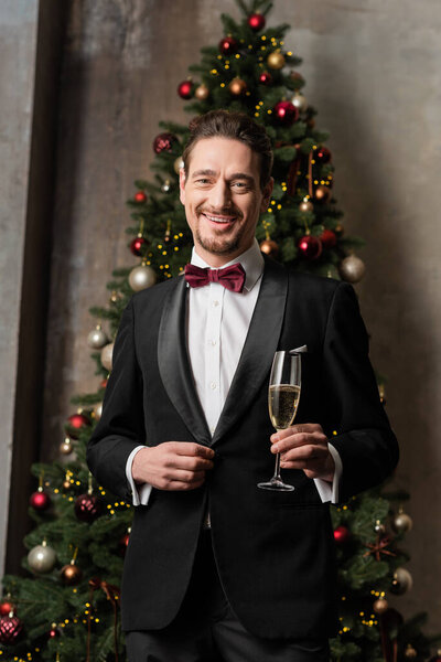 cheerful gentleman in suit with bow tie holding champagne glass near decorated Christmas tree