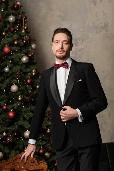 wealthy gentleman in formal attire with bow tie looking at camera near decorated Christmas tree