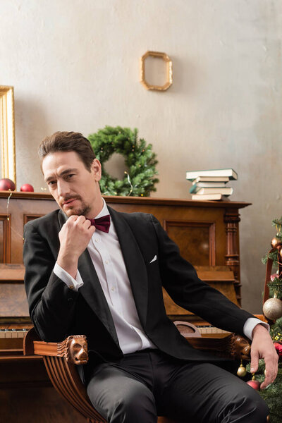 elegant gentleman in tuxedo with bow tie sitting near piano with books and Christmas baubles