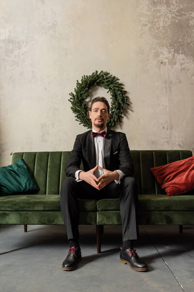 well-dressed gentleman with beard wearing tuxedo with bow tie sitting on sofa near Christmas wreath