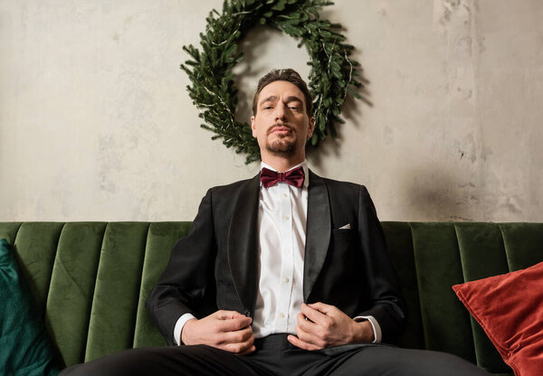 wealthy gentleman with beard wearing tuxedo with bow tie sitting on sofa near Christmas wreath