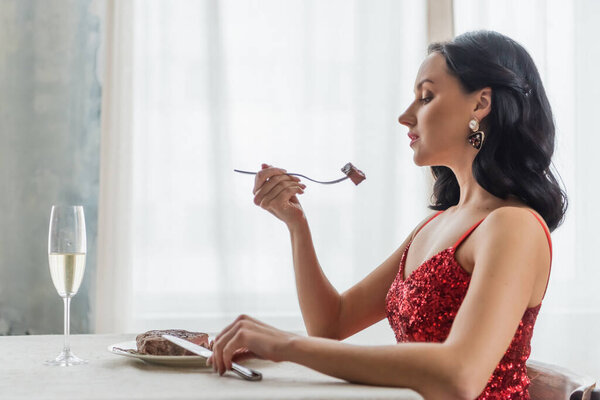 elegant woman in red dress sitting at dining table with glass of champagne and eating beef steak