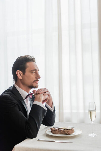 красивый джентльмен в смокинге сидит за обеденным столом с бокалом шампанского и бифштексом на тарелке