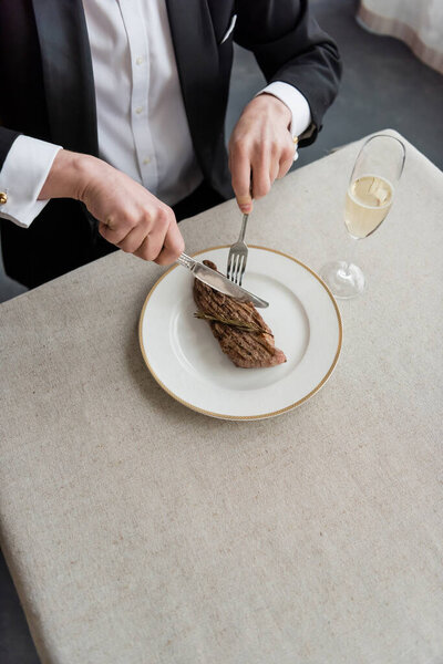 top view of wealthy man in tuxedo cutting delicious beef steak on plate near glass of champagne