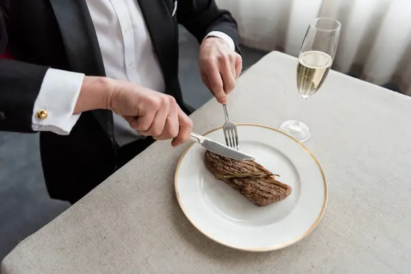 overhead view of wealthy man in tuxedo cutting delicious beef steak on plate near glass of champagne
