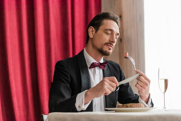 well-dressed man in tuxedo enjoying taste of beef steak near champagne in glass on dining table